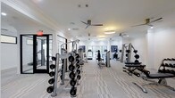 The fitness center is a thoughtfully designed workout space that balances functionality with refined aesthetics. The interior features crisp white walls adorned with decorative athletic-themed artwork, complemented by crown molding that adds a polished architectural detail. Underfoot, patterned low-pile carpeting runs throughout, providing both comfort and durability during workouts.

The space is generously proportioned, accommodating a wide array of equipment without feeling crowded. A dedicated free weights section includes a fully stocked dumbbell rack ranging from light to heavy loads, ideal for strength training routines. Across the floor, cable and resistance machines from Hoist are arranged to allow easy circulation between stations. Toward the back of the room, a row of cardio machines — including treadmills and ellipticals — faces large windows that invite natural light and provide outdoor views, creating an energizing atmosphere during exercise.

Overhead, multiple modern ceiling fans with sleek metallic finishes promote air circulation throughout the space. Recessed lighting and a circular pendant fixture provide even, well-distributed illumination. A glass-paneled entry door enhances the open feel and connects the fitness center to adjacent areas of the building.
