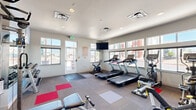 Located at 5923 Tutt Blvd in Colorado Springs, Colorado, this well-equipped fitness center offers a thoughtfully organized workout space on a single floor. The room features neutral-toned walls, recessed and flush-mount ceiling lighting, and a clean white ceiling that keeps the atmosphere bright and energizing.

Large windows line multiple walls, flooding the space with abundant natural light and offering views of the surrounding neighborhood. A wall-mounted flat-screen television provides entertainment during workouts, while full-length mirrors along one wall visually expand the room and allow users to monitor their form.

The cardio section is well-stocked with multiple treadmills, an elliptical trainer, a stationary upright bike, and a cycling bike, all positioned to take advantage of the natural light from the windows. The strength training area features a multi-station cable machine, a dedicated weight bench, and a fully stocked dumbbell rack with a broad range of free weights. A designated stretching and floor exercise zone is marked by colorful interlocking floor tiles — red, gray, and white — set against the room's neutral carpeted flooring.

The space is accessed via a glass-paneled entry door and maintains a clean, modern aesthetic throughout. With a comprehensive mix of cardio and strength equipment, this fitness center provides a functional and well-lit environment suited to a wide variety of workout routines.