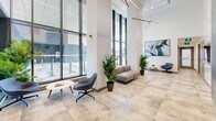 This contemporary building lobby makes an immediate impression with its dramatically high ceilings and expansive floor-to-ceiling glass curtain walls that flood the interior with abundant natural light and offer open views to the outdoors. The floor is finished in large-format stone-look porcelain tiles in warm neutral tones, lending the space a refined, professional character.

Overhead, a striking sculptural pendant light fixture with warm wood-toned arms serves as a focal point, complemented by recessed ceiling lighting that ensures even illumination throughout. The white walls provide a clean, crisp backdrop, with curated abstract artwork adding visual interest at key points.

Multiple seating arrangements are thoughtfully distributed across the lobby, including a modular sofa grouping, paired lounge chairs with a round side table, and individual accent chairs — all in cool grey tones that harmonize with the overall palette. Lush tropical potted plants are placed throughout, introducing organic warmth and vitality to the space.

A vestibule entry with glass doors provides secure and weather-controlled access from the exterior, while additional doors lead to corridors beyond. A wall-mounted display panel and marble-effect wall cladding near the elevator area add further architectural detail. The overall ambiance is welcoming, polished, and professional — well-suited to a modern commercial or mixed-use building.