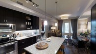 This single-floor space showcases a thoughtfully designed open-concept layout that integrates the kitchen, dining, and living areas into one cohesive environment. The kitchen is a standout feature, equipped with dark espresso cabinetry that contrasts elegantly against a light granite countertop island, which serves as a natural focal point for the space. A classic subway tile backsplash, an undermount sink, and track lighting overhead add functional and aesthetic detail. Three pendant lights hang above the island, providing warm, layered illumination that complements the space's modern ambiance.

The open layout flows naturally into the dining area, where a glass-top table with industrial-style stools continues the contemporary aesthetic. Further into the space, the living area features a seating arrangement anchored by a neutral-toned armchair, a decorative console table with a table lamp, and floor-length curtains that frame a glass door leading to what appears to be an exterior space, allowing natural light to filter in. The hardwood-style flooring runs throughout, unifying the entire open-plan space and reinforcing the clean, modern character of the home.