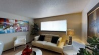 This single-floor space offers a welcoming and well-appointed living area designed with comfort and functionality in mind. The living room serves as the centerpiece of the space, featuring warm, neutral-toned walls that create a cozy and inviting ambiance. A textured popcorn ceiling adds a classic architectural character to the room, while a wide horizontal window fitted with white horizontal blinds allows for an ample influx of natural light, brightening the interior throughout the day.

The living area is furnished with a sleek, cream-colored leather sofa accented with dark throw pillows, complemented by matching white armchairs that contribute to the room's clean, contemporary aesthetic. A stylish table lamp with a metallic base provides warm accent lighting, adding to the room's inviting atmosphere. Decorative touches, including a bold abstract wall painting and a vibrant red decorative vase, inject personality and visual interest into the space. A lush indoor plant adds a natural, refreshing element to the corner of the room. The flooring features wall-to-wall carpeting that enhances the overall warmth and comfort of the living area, making this a thoughtfully designed and cohesive single-floor space.