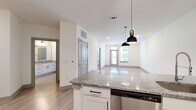 This well-appointed single-floor residence offers a thoughtfully designed layout encompassing one bedroom, one bathroom, a combined kitchen and living area, and a dedicated closet. The open-concept kitchen and living room serves as the heart of the space, featuring a large granite-topped island with an undermount sink and stylish pendant lighting overhead. White shaker cabinetry lines the kitchen walls, complemented by a full suite of stainless steel appliances including a refrigerator with ice maker, electric range, and built-in microwave. Light wood-style plank flooring extends throughout the main living areas, reflecting natural light that enters through large windows and a glass-paneled exterior door leading to a balcony with neighborhood views. The living area is generously proportioned with recessed lighting and a clean, neutral color palette. The bedroom is carpeted for comfort, offering two large windows that invite abundant natural light and outdoor views. A dedicated closet provides convenient storage. The bathroom is finished with the same granite countertop found in the kitchen, set atop a white vanity with modern hardware, and includes a full tiled tub-shower combination with a curved rod and recessed lighting.