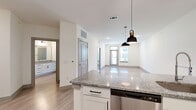 This well-appointed single-floor residence offers a thoughtfully designed layout encompassing one bedroom, one bathroom, a combined kitchen and living area, and a dedicated closet. The open-concept kitchen and living room serves as the heart of the space, featuring a large granite-topped island with an undermount sink and stylish pendant lighting overhead. White shaker cabinetry lines the kitchen walls, complemented by a full suite of stainless steel appliances including a refrigerator with ice maker, electric range, and built-in microwave. Light wood-style plank flooring extends throughout the main living areas, reflecting natural light that enters through large windows and a glass-paneled exterior door leading to a balcony with neighborhood views. The living area is generously proportioned with recessed lighting and a clean, neutral color palette. The bedroom is carpeted for comfort, offering two large windows that invite abundant natural light and outdoor views. A dedicated closet provides convenient storage. The bathroom is finished with the same granite countertop found in the kitchen, set atop a white vanity with modern hardware, and includes a full tiled tub-shower combination with a curved rod and recessed lighting.