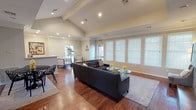 This single-floor space offers an open, welcoming layout highlighted by soaring vaulted ceilings with exposed beams and recessed lighting that create a bright, airy atmosphere. Rich hardwood flooring runs throughout, adding warmth and continuity to the interior. The main living area is furnished with a dark leather sectional sofa, complemented by a glass-top coffee table and a patterned area rug, creating an inviting seating arrangement. Yellow floral accents provide pops of color against the neutral palette of warm white and beige walls. A row of large windows dressed with white horizontal blinds lines one wall, allowing generous natural light to fill the space while maintaining privacy. Additional seating is available in a separate lounge area toward the rear, featuring armchairs and a side table alongside a potted plant that introduces a natural element. A counter or bar-height surface is visible to one side, suggesting a supplementary utility or serving area. The overall ambiance is clean, modern, and functional, with thoughtful furnishings that balance comfort and style throughout the space.