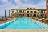 Located at 320 Broad Street in the heart of Charleston, South Carolina, this outdoor terrace offers a thoughtfully designed open-air environment that blends relaxed sophistication with functional amenity space. The terrace is finished with large-format stone pavers in warm neutral tones, providing a clean, cohesive foundation throughout the space.

Generous seating areas are distributed across the terrace, featuring wicker and rattan lounge furniture arranged around low coffee tables, ideal for casual gatherings. A separate dining zone is outfitted with sleek, modern tables and woven chairs, accommodating group meals in an al fresco setting. Large concrete planters containing ornamental trees are positioned strategically across the terrace, providing natural greenery and subtle spatial definition without obstructing the open feel.

An outdoor kitchen station, built into a classic brick surround, includes a stainless steel grill and prep area, making the space well-suited for outdoor entertaining. Glass-paneled entry doors connect the terrace to an adjacent interior space, flanked by traditional wall-mounted lanterns that complement Charleston's architectural character.

The terrace benefits from unobstructed views toward the waterfront, with open sky and coastal scenery defining the horizon, creating a bright and airy atmosphere throughout the day.