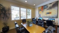 This business center is designed to provide a functional and comfortable professional environment for productivity and collaboration. The single-floor space features warm, neutral-toned walls accented by elegant crown molding, creating a polished and welcoming atmosphere. A central wooden conference table is surrounded by plush leather executive chairs, offering an ideal setting for meetings or focused work sessions. Along one wall, a dedicated workstation area is equipped with multiple desktop computers, providing convenient access to technology and connectivity. Large windows with white horizontal blinds line one side of the room, allowing generous natural light to fill the space while offering a view of the surrounding outdoor area. Recessed ceiling lighting further brightens the interior for consistent illumination throughout the day. A vibrant large-scale abstract artwork adorns one wall, adding a splash of color and creative energy to the otherwise professional setting. The carpeted flooring and thoughtfully arranged furnishings contribute to an overall ambiance that is both productive and inviting, making this business center a well-rounded resource for professional needs.