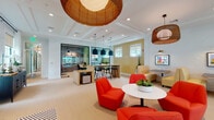 Located on Garden Catalina Circle in Lake Worth, Florida, this single-story community clubhouse is thoughtfully designed to accommodate a variety of social and functional needs. The space is organized into several distinct zones that transition seamlessly from one to the next.

The grand entrance features soaring double-height ceilings, shiplap wall detailing, and a striking arched transom window above French doors that floods the foyer with natural light. Herringbone-patterned wood-look flooring anchors this welcoming area, complemented by lush tropical greenery and a plaid upholstered bench.

The main lounge area continues the elevated aesthetic with a dramatic equine mural serving as a bold focal point above a communal dining table with wishbone chairs. Adjacent seating areas offer leather sofas, plaid ottomans, and woven pendant lighting beneath the towering ceiling.

A separate, brighter wing transitions into a casual coworking and lounge space, featuring coffered ceilings, woven rattan pendants, bold red lounge chairs, a bar-height work counter with stools, glass-enclosed private rooms, built-in cabinetry with a beverage station, and wall-mounted television. Large steel-framed windows throughout provide views of the surrounding Florida landscape, reinforcing the space's open, airy ambiance.