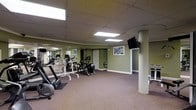 This single-floor fitness center offers a practical and functional space designed to support a well-rounded workout routine. The room features a selection of cardio equipment, including treadmills, an elliptical, and a rowing machine, alongside strength training apparatus, providing users with a comprehensive range of exercise options. A wall-mounted flat-screen television is positioned on a structural column near the center of the room, allowing for entertainment during workouts. A large mirrored wall along one side of the room visually expands the space and assists users in monitoring their form during exercise. The walls are painted in a muted sage green tone, contributing to a calm and focused atmosphere. Recessed panel lighting in the drop ceiling, supplemented by skylight-style flush panels, ensures the space is well-lit throughout. Framed artwork adorns the walls, adding a subtle decorative touch to the otherwise utilitarian environment. Carpeted flooring with rubberized exercise mats beneath the equipment provides comfort and stability. A doorway leads to an adjacent area, and a small water station is visible along the wall, adding to the practical amenities of this well-appointed fitness facility.