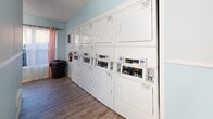 Located in Augusta, Georgia, this shared laundry facility is designed to provide a practical and comfortable experience for its users. The single-floor space is finished with durable wood-look vinyl flooring throughout, offering both easy maintenance and a clean aesthetic. The walls are painted in a soft, light blue tone, creating a calm and welcoming atmosphere brightened by overhead fluorescent lighting.

The facility is well-stocked with multiple commercial-grade top-load washing machines and a bank of stacked dryers lining the walls, all featuring digital control panels for ease of use. The arrangement allows for efficient use of the space while keeping the floor area open and accessible.

A dedicated folding station with a large white table and seating provides a convenient area for sorting and folding laundry. A curtained window allows natural light to filter in, adding to the overall brightness of the room. A fire extinguisher and waste bin are thoughtfully positioned for safety and cleanliness. A pass-through door provides additional access and ventilation. The layout is straightforward and functional, making this laundry room a practical and well-maintained shared amenity.