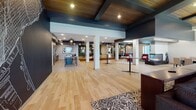 Located at 201 Clover St in Duluth, Minnesota, this thoughtfully designed single-floor amenity space offers a diverse range of gathering areas tailored for both relaxation and socializing. The open main hall features warm light hardwood flooring, bold navy ceiling beams with wood inlays, and recessed lighting that create an inviting, modern atmosphere throughout.

The fully equipped kitchen showcases warm wood cabinetry, stainless steel appliances, a deep green tile backsplash, and a large dark-topped island with bar seating. Distinctive sculptural pendant lights add a dramatic focal point overhead, while patterned tile flooring grounds the space with texture and visual interest.

A dedicated lounge and seating area features wraparound tufted leather banquette seating, round bistro tables, and accent chairs set against a striking hexagonal tile feature wall. Decorative lattice-panel dividers provide elegant spatial definition throughout.

A sleek beverage and coffee bar station is outfitted with modern dispensing equipment, matte black cabinetry, glossy black vertical subway tile, and open wood shelving — ideal for daily use.

The formal dining area accommodates a large party around a substantial dark wood table beneath statement drum pendants. Floor-to-ceiling windows flood the room with natural light and provide direct views and access to an outdoor patio courtyard, seamlessly connecting interior and exterior living.