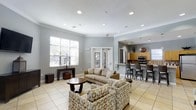 This single-floor space offers a thoughtfully designed open-concept layout that seamlessly blends a comfortable lounge area with a functional kitchen, making it ideal for social gatherings and everyday use. The lounge is furnished with a neutral-toned sofa and patterned accent chairs arranged around a dark wood coffee table, creating an inviting setting for relaxation or conversation. Light tile flooring runs throughout, contributing to a clean and airy aesthetic. The room is finished with crown molding and recessed lighting in a smooth white ceiling, adding a refined architectural touch. Large windows fitted with white plantation shutters allow generous natural light to fill the space. French doors provide an additional connection to an adjacent area, enhancing the open flow of the layout. The kitchen, visible from the lounge through an open pass-through, features warm wood cabinetry, stainless steel appliances, a tiled backsplash, and a raised bar counter lined with dark bar stools. A pendant light hangs above the counter, adding a decorative focal point. The overall ambiance is bright, neutral, and welcoming, with a balanced mix of functional and comfortable design elements throughout.