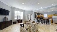 This single-floor space offers a thoughtfully designed open-concept layout that seamlessly blends a comfortable lounge area with a functional kitchen, making it ideal for social gatherings and everyday use. The lounge is furnished with a neutral-toned sofa and patterned accent chairs arranged around a dark wood coffee table, creating an inviting setting for relaxation or conversation. Light tile flooring runs throughout, contributing to a clean and airy aesthetic. The room is finished with crown molding and recessed lighting in a smooth white ceiling, adding a refined architectural touch. Large windows fitted with white plantation shutters allow generous natural light to fill the space. French doors provide an additional connection to an adjacent area, enhancing the open flow of the layout. The kitchen, visible from the lounge through an open pass-through, features warm wood cabinetry, stainless steel appliances, a tiled backsplash, and a raised bar counter lined with dark bar stools. A pendant light hangs above the counter, adding a decorative focal point. The overall ambiance is bright, neutral, and welcoming, with a balanced mix of functional and comfortable design elements throughout.