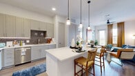 This single-floor, 2-bedroom, 2-bathroom residence offers a well-organized layout with modern finishes and a cohesive design throughout. The living room features light wood-style flooring, a ceiling fan, large windows with floor-to-ceiling drapes that invite natural light, and ample space for comfortable seating. The kitchen is equipped with stainless steel appliances including a full-size refrigerator, range, microwave, and dishwasher, complemented by gray shaker-style cabinetry, white quartz countertops, a decorative geometric tile backsplash, and a kitchen island for additional prep space. The primary bedroom provides a generously sized retreat with plush carpeting and direct access to its en-suite bathroom, which features a dual-sink vanity with backlit mirrors, wood-look flooring, a soaking tub, a glass-enclosed shower with tile surround, and a walk-in closet with built-in shelving and hanging rods. The second bedroom offers carpeted flooring and a large window with outdoor views. The second bathroom also includes a single vanity with a backlit mirror and tub-shower combination. Both bedrooms are served by dedicated closets, providing excellent storage throughout the residence.
