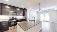 This well-designed one-bedroom, two-bathroom residence offers an efficient and thoughtfully laid-out single-floor plan. The open-concept kitchen and living area serves as the heart of the home, anchored by a large central island with quartz countertops and an undermount sink with a brushed gold faucet. Pendant lighting with glass jar-style fixtures adds a contemporary touch overhead. Rich, dark espresso cabinetry extends floor-to-ceiling, providing ample storage, while a full suite of stainless steel appliances—including a refrigerator, electric range, and built-in microwave—completes the modern kitchen. Light hardwood-style flooring runs throughout the entire unit, lending warmth and visual continuity. The living area is bright and airy, featuring a ceiling fan and French doors that invite natural light and provide outdoor access. The spacious primary bedroom includes a well-organized built-in closet system with hanging rods and open shelving. Both bathrooms are finished with dark cabinetry vanities, quartz countertops, and brushed nickel hardware. One bathroom features a full soaking tub with white subway tile surround, while the other offers a clean, functional layout. The overall aesthetic is clean and contemporary throughout.