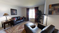 This two-floor residence offers a thoughtfully arranged living space designed for comfort and everyday functionality. The living room serves as a welcoming focal point, featuring dark leather seating — including a sofa and armchair — arranged atop a richly patterned area rug that adds warmth and visual depth to the neutral-toned carpeted floor. A large window dressed with layered, earth-toned curtains in deep reds and warm neutrals allows natural light to filter into the room while providing privacy, and views of greenery outdoors can be glimpsed through the glass. A tall fiddle-leaf fig plant in the corner adds a natural, organic element to the space, complementing the understated wall décor, including a framed botanical artwork. Clean white walls and a smooth ceiling keep the aesthetic simple and uncluttered, allowing the furnishings and accents to take center stage. A side table anchors one end of the sofa, providing a functional surface. The overall ambiance of the living area is warm and relaxed, with a classic residential character. Spanning two floors, the home offers ample room to accommodate a variety of living arrangements, with the upper and lower levels providing distinct zones for rest, recreation, and daily living.
