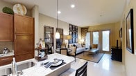 This single-floor Dallas residence showcases a thoughtfully designed open-concept layout that unifies the kitchen, dining, and living areas into one cohesive, flowing space. The kitchen features a striking marble-patterned countertop that serves as both a functional workspace and a casual dining surface, anchored by sleek pendant lighting that adds a warm, inviting glow. The dining area sits adjacent, furnished with dark-framed chairs with nail-head trim detailing, positioned beneath a coordinating pendant light that reinforces the space's refined aesthetic. A pair of large framed mirrors with geometric black metalwork amplifies the sense of depth and light in the room. The living area extends naturally from the dining space, centered around a neutral-toned sofa and complementary seating arranged on a patterned area rug over light-toned tile flooring. A gallery wall of botanical prints adds visual interest, while floor-to-ceiling sliding glass doors flood the space with natural light and offer views of the surrounding greenery. Recessed ceiling lighting throughout ensures the interior remains bright and well-illuminated, contributing to the space's clean, contemporary ambiance.