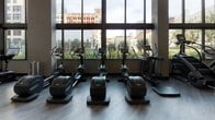 This two-floor fitness facility is designed with a contemporary aesthetic, combining functionality with a polished, modern finish. The interior features wood-look flooring, linear LED lighting, and a neutral color palette of white and dark gray accents that lend a professional, high-end atmosphere throughout.

The strength training area is well-equipped with a full rack of dumbbells, adjustable benches, cable machines, and weight equipment arranged across padded mat flooring. Floor-to-ceiling mirrors amplify the sense of space and allow for proper form monitoring. A dedicated functional training zone features a RaX rig system stocked with kettlebells, medicine balls, battle ropes, TRX suspension trainers, and a heavy punching bag, all positioned on rubberized tile flooring ideal for dynamic movement.

The cardio floor is situated adjacent to an expansive wall of floor-to-ceiling windows that frame views of the surrounding urban landscape and landscaped terrace below, filling the space with natural light. Equipment includes treadmills, ellipticals, and water rowing machines paired with a mounted display screen. A water fountain and bottle-filling station provide convenient hydration access. Artistic wall decor, including a striking figure mural on the upper level, adds a creative touch to the energetic environment.