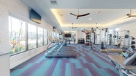 This modern fitness center is designed to support a comprehensive workout experience within a single, open-plan floor. The space is defined by its striking teal and purple patterned carpet tiles, which add a vibrant, energetic ambiance throughout. A tray ceiling with warm recessed and cove lighting creates a polished architectural finish, while multiple ceiling fans ensure consistent air circulation across the floor.

The cardio zone runs along a wall of large windows, housing a row of treadmills, an elliptical, and a stair climber. These windows offer abundant natural light and direct views of an outdoor pool and landscaped surroundings, enhancing the workout environment. Mounted flat-screen TVs are positioned at eye level along this wall for added convenience.

The strength training area occupies the opposite side of the space and is thoroughly equipped with a multi-station cable machine system, weight benches, and a well-stocked dumbbell rack. A dedicated functional training section includes medicine ball racks, TRX suspension trainers, and resistance accessories. Full-length mirrors in the strength zone allow users to monitor their form. A textured stone accent wall adds a design element that grounds the space visually. The fitness center is accessed through a glass-panel door and benefits from both natural light and thoughtfully designed artificial lighting throughout.