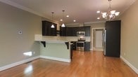 This three-floor home showcases a thoughtfully designed open-concept main living area that seamlessly connects the kitchen and living spaces. The kitchen is a standout feature, appointed with rich, dark espresso-toned cabinetry that provides a striking contrast against the light sage-green walls and warm honey-toned hardwood floors that run throughout. A peninsula with granite countertops and decorative bracket supports offers additional prep space and casual seating, making it ideal for entertaining.

Stainless steel appliances, including a range and built-in microwave, are neatly integrated into the cabinetry. Overhead, a combination of recessed lighting and pendant lights provides layered illumination, while crown molding along the ceiling adds a refined architectural detail. A partial view of an adjacent hallway hints at additional rooms and functional spaces beyond.

The open layout promotes an airy, connected feel across the main level, while the home's three-floor layout suggests ample space for living, sleeping, and additional utility areas on the remaining levels. The overall aesthetic balances classic design elements with modern finishes.