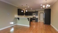 This three-floor home showcases a thoughtfully designed open-concept main living area that seamlessly connects the kitchen and living spaces. The kitchen is a standout feature, appointed with rich, dark espresso-toned cabinetry that provides a striking contrast against the light sage-green walls and warm honey-toned hardwood floors that run throughout. A peninsula with granite countertops and decorative bracket supports offers additional prep space and casual seating, making it ideal for entertaining.

Stainless steel appliances, including a range and built-in microwave, are neatly integrated into the cabinetry. Overhead, a combination of recessed lighting and pendant lights provides layered illumination, while crown molding along the ceiling adds a refined architectural detail. A partial view of an adjacent hallway hints at additional rooms and functional spaces beyond.

The open layout promotes an airy, connected feel across the main level, while the home's three-floor layout suggests ample space for living, sleeping, and additional utility areas on the remaining levels. The overall aesthetic balances classic design elements with modern finishes.