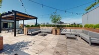 Located at 1120 Frankford Avenue in Philadelphia's Fishtown neighborhood, this expansive rooftop deck offers a thoughtfully designed outdoor living experience with striking views of the Philadelphia skyline. The space is finished with wide-plank wood decking throughout and is organized into several distinct functional zones.

A bold steel pergola with wood-paneled ceiling accents and ceiling fans anchors a covered dining area, furnished with multiple tables and chairs — ideal for al fresco gatherings. String lights are strung across the open-air portions of the deck, adding ambiance for evening use.

A generously sized lounge zone features modular sectional sofas with neutral cushions, complemented by large raised wooden planters filled with manicured boxwood and ornamental trees that provide both privacy and greenery. A rectangular fire pit serves as a focal point in this area, creating a comfortable setting for year-round enjoyment.

An outdoor kitchen station includes a built-in grill, prep surfaces, and a bar-height island with seating, offering a fully functional cooking and entertaining area. The surrounding cedar-clad privacy walls add warmth and definition to the space.

Glass-panel railings along the perimeter maintain unobstructed panoramic views of the Philadelphia skyline and the surrounding Fishtown and Kensington neighborhoods, making this rooftop a standout amenity.