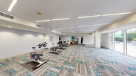Located at 700 SW 78th Ave in Plantation, FL, this well-appointed fitness studio offers a thoughtfully designed single-floor space ideal for a variety of workout styles. The room is finished with a distinctive patterned carpet in grey and teal tones, contributing to a clean, contemporary aesthetic throughout.

The studio is equipped with a row of cycling bikes positioned toward the center of the room, providing ample space for spin classes or individual cardio sessions. A dedicated functional training zone features a standing rack stocked with stability balls in multiple sizes, medicine balls, resistance bands, and rolled exercise mats — offering everything needed for strength and flexibility training. A heavy punching bag is suspended from the ceiling, adding a combat fitness element to the space.

Linear LED lighting is recessed into the white ceiling, delivering bright, even illumination across the entire floor. A wall-mounted flat-screen television is positioned near a storage console, supporting instructor-led or on-demand workout sessions. Large sliding glass doors along one wall flood the space with natural light and provide direct access to an outdoor area, creating an open, airy atmosphere. A glass-paneled interior entrance is etched with signage, reinforcing the studio's dedicated purpose. The combination of modern finishes, diverse equipment, and abundant light makes this a highly functional and inviting fitness environment.