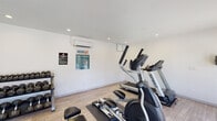 The Seventeen805 Fitness Center is a clean, modern facility designed to provide residents with a comprehensive workout experience. The space features wood-look tile flooring throughout and is finished with crisp white walls and recessed lighting that keeps the environment bright and welcoming.

The cardio section is equipped with Spirit-brand treadmills, an elliptical trainer, and a rowing machine, all arranged to maximize floor space and ease of movement. A dedicated free weights area features two multi-tiered dumbbell racks with a full range of weights, complemented by exercise mats for stretching or floor work. Full-length wall mirrors run along one side of the room, enhancing the sense of openness and allowing users to monitor their form during workouts.

Climate control is provided via wall-mounted mini-split units, ensuring year-round comfort. Glass panel doors line one wall, allowing natural light to filter in while offering direct access to the outdoor pool and patio area, where lounge seating and a covered pergola are visible. Posted fitness center rules and policy notices keep the space organized and informative for all users. The overall ambiance is clean, functional, and community-focused, making it a practical amenity for everyday fitness routines.