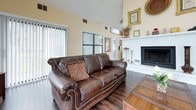 Located at 397 Brixton Drive in Virginia Beach, Virginia, this single-story home delivers a compelling blend of comfort and architectural character. The living area is a standout, boasting dramatically vaulted ceilings with exposed wooden beams and a sweeping wall of floor-to-ceiling triangular windows that flood the space with natural light and offer views of the surrounding greenery. Dark hardwood flooring runs throughout, anchoring the warm, neutral color palette of the interior. A classic white brick wood-burning fireplace serves as a focal point, adding charm and a sense of coziness to the living space. The kitchen is thoughtfully designed with warm honey-toned cabinetry, dark granite-style countertops, and a full suite of appliances including a black refrigerator, microwave, and stainless steel dishwasher. A built-in desk area within the kitchen provides a functional workspace seamlessly integrated into the cabinetry. Large windows in the kitchen invite additional natural light, further enhancing the open and airy feel of the home. The single-floor layout ensures accessibility and ease of movement throughout, while the cohesive design elements create a welcoming and well-organized living environment throughout the entire space.