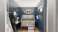 Located at 915 West Grape Street in San Diego, CA, this purpose-built pet grooming space is a compact, single-room facility designed with both functionality and style in mind. The centerpiece of the room is a professional-grade stainless steel dog wash tub, complete with step-up access for pets and a retractable blue hose for convenient rinsing. Rich navy blue walls give the space a polished, boutique feel, while warm wood-look flooring adds a touch of comfort underfoot. Decorative rugs flank the tub area, and gold-framed pet portraits arranged on the back wall lend a charming, curated aesthetic. Wall-mounted sconce lighting provides focused illumination throughout. A small side shelf offers workspace for grooming tools and supplies, while hooks along the wall provide convenient storage for aprons and accessories. The space is accessed through a heavy-duty door adjacent to a utility corridor, reflecting the building's robust construction with exposed concrete masonry and industrial-grade infrastructure. The broader building context includes a parking structure with visible overhead ductwork, underscoring the mixed-use, urban nature of this San Diego property. Overall, this grooming station strikes a balance between professional utility and thoughtful design.