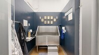 Located at 915 West Grape Street in San Diego, CA, this purpose-built pet grooming space is a compact, single-room facility designed with both functionality and style in mind. The centerpiece of the room is a professional-grade stainless steel dog wash tub, complete with step-up access for pets and a retractable blue hose for convenient rinsing. Rich navy blue walls give the space a polished, boutique feel, while warm wood-look flooring adds a touch of comfort underfoot. Decorative rugs flank the tub area, and gold-framed pet portraits arranged on the back wall lend a charming, curated aesthetic. Wall-mounted sconce lighting provides focused illumination throughout. A small side shelf offers workspace for grooming tools and supplies, while hooks along the wall provide convenient storage for aprons and accessories. The space is accessed through a heavy-duty door adjacent to a utility corridor, reflecting the building's robust construction with exposed concrete masonry and industrial-grade infrastructure. The broader building context includes a parking structure with visible overhead ductwork, underscoring the mixed-use, urban nature of this San Diego property. Overall, this grooming station strikes a balance between professional utility and thoughtful design.