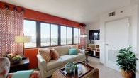 This single-floor residence is situated on an upper level of a high-rise building, offering sweeping cityscape and treetop views through large, dark-framed windows throughout. The living room serves as the centerpiece of the unit, featuring a bold terra-cotta accent wall that commands attention alongside expansive windows that flood the space with natural light. Neutral carpet, a comfortable sofa, woven coffee table, and an eclectic multi-drawer media console contribute to the room's colorful, layered character.

Adjacent to the living area, the dining space is defined by soft aqua-painted walls, a pendant light over an oval dining table, and a decorative round mirror — creating a calm, welcoming atmosphere for meals. The compact kitchen features white cabinetry, light countertops, and full appliances including a gas range and dishwasher, with green-painted walls adding warmth.

The bedrooms are carpeted and well-proportioned, each offering access to outdoor views through individual windows. The primary bedroom includes a large closet with built-in shelving, providing ample storage. A carpeted hallway connects the rooms, lending a quiet, private feel to the sleeping quarters. Overall, the unit balances vibrant design choices with comfortable, functional living spaces.