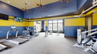Located at 10800 McCombs St, this fitness center is a thoughtfully designed exercise space that offers a comprehensive workout environment. The room is immediately distinguished by its bold, two-tone color scheme of deep navy blue and vibrant yellow, which energizes the space and gives it a dynamic, motivating atmosphere. Elevated ceilings with decorative ceiling fans enhance the sense of openness and promote air circulation throughout the room.

The fitness center is divided into functional zones. The cardio area features multiple treadmills and elliptical machines, each equipped with individual monitors and positioned in front of wall-mounted flat-screen televisions. The strength training section includes a variety of cable and resistance machines, a weight bench, and a dumbbell rack. A dedicated free weight and stretching area provides ample open floor space, complemented by medicine balls and exercise mats.

Large floor-to-ceiling windows along one wall flood the space with natural light and offer direct views of an outdoor pool, creating an inviting and refreshing workout backdrop. A separate nook features a textured accent wall, a mounted TV, and additional free weights, offering a more private training area. The carpeted flooring, recessed lighting, and framed artwork complete a polished and functional fitness facility.