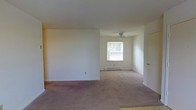 This single-floor residence offers a straightforward and functional layout designed for comfortable everyday living. The interior is characterized by neutral white walls and soft, wall-to-wall carpeting in a warm taupe tone that runs throughout the main living areas, creating a cohesive and understated aesthetic.

The open living space is generously proportioned, allowing for flexible furniture arrangements and a sense of easy flow between areas. A partial wall divides the space, adding subtle definition without sacrificing the open feel. Natural light filters in through a window that looks out onto surrounding greenery, brightening the interior and providing a pleasant connection to the outdoors.

A ceiling fan is installed in the adjoining area, contributing to year-round comfort and air circulation. Baseboard heating runs along the perimeter, ensuring efficient warmth throughout the cooler months. A standard interior door provides access to additional rooms, suggesting a practical and well-organized floor plan.

The overall ambiance is clean, quiet, and move-in ready, with a simple palette that lends itself well to personalization. The space is well-suited for those seeking a low-maintenance, single-level living environment with a calm and neutral interior character.