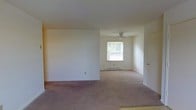 This single-floor residence offers a straightforward and functional layout designed for comfortable everyday living. The interior is characterized by neutral white walls and soft, wall-to-wall carpeting in a warm taupe tone that runs throughout the main living areas, creating a cohesive and understated aesthetic.

The open living space is generously proportioned, allowing for flexible furniture arrangements and a sense of easy flow between areas. A partial wall divides the space, adding subtle definition without sacrificing the open feel. Natural light filters in through a window that looks out onto surrounding greenery, brightening the interior and providing a pleasant connection to the outdoors.

A ceiling fan is installed in the adjoining area, contributing to year-round comfort and air circulation. Baseboard heating runs along the perimeter, ensuring efficient warmth throughout the cooler months. A standard interior door provides access to additional rooms, suggesting a practical and well-organized floor plan.

The overall ambiance is clean, quiet, and move-in ready, with a simple palette that lends itself well to personalization. The space is well-suited for those seeking a low-maintenance, single-level living environment with a calm and neutral interior character.