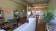 This single-floor space showcases an industrial aesthetic defined by exposed ductwork running along the ceiling, complemented by sleek track lighting that illuminates the open-concept interior. The living area is anchored by a white sofa and a low-profile coffee table set atop a textured area rug, with a glass-door display cabinet adding a decorative accent nearby. Dark hardwood flooring flows continuously throughout, grounding the space with warmth against the cool industrial elements above.

The kitchen is fully integrated into the living space, featuring stainless steel appliances including a refrigerator, gas range, and microwave. Warm wood cabinetry lines the upper walls, providing generous storage, while a raised breakfast bar with bar seating serves as a natural dividing element between the kitchen and living zones. A pendant light hangs over the bar area, adding a focused point of illumination.

The elevated cabinetry arrangement and high ceilings contribute to a sense of vertical spaciousness, while the open floor plan creates a fluid, connected feel throughout the entire living and kitchen area. The overall ambiance blends urban industrial character with functional, everyday comfort.