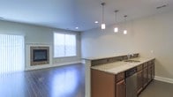 This two-story space offers a thoughtfully designed open-concept layout that seamlessly connects the kitchen and living area. The kitchen is appointed with warm wood cabinetry and elegant granite countertops that extend across a peninsula, providing both workspace and a casual dining option. Three pendant lights hang above the peninsula, offering focused task lighting while adding a polished aesthetic touch. Stainless steel appliances complete the kitchen's functional design.

The adjoining living area features rich hardwood flooring that runs throughout, lending warmth and continuity to the space. A tile-surround fireplace serves as the focal point of the living room, providing both visual interest and a cozy gathering spot. A window fitted with blinds allows natural light to filter into the room while maintaining privacy. Neutral-toned walls and recessed ceiling lighting throughout contribute to a bright, clean atmosphere that complements the space's modern yet inviting character. The two-floor layout suggests additional living or sleeping accommodations on the upper level, expanding the overall functionality of the home.