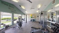 This fitness center is thoughtfully designed to support a full range of workout routines across two distinct zones. The main strength training area features vaulted ceilings that lend an open, airy feel, accentuated by soft sage-green accent walls and the motivational phrase "Think Strong, Be Strong, Finish Strong" displayed prominently. Large windows line the perimeter, flooding the space with natural light and offering views of the surrounding greenery. Ceiling fans provide additional ventilation throughout.

The strength zone is comprehensively outfitted with a multi-station cable machine, a full rack of dumbbells, an adjustable weight bench, and a variety of selectorized machines for targeted muscle training. A dedicated storage rack holds stability balls and medicine balls of varying sizes, supporting functional and core training. Full-length mirrors along one wall enhance the sense of space while allowing users to monitor their form.

A separate cardio room houses multiple treadmills and an elliptical machine, each equipped with individual display consoles. This area benefits from its own set of windows overlooking outdoor landscaping, along with a wall-mounted television for entertainment during workouts. Durable carpet tile flooring runs throughout the entire facility, and the space is accessible via a connecting hallway.