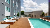 This amenity space combines an outdoor pool area and a fully equipped fitness center, offering residents a well-rounded recreational experience. The outdoor pool features crystal-clear turquoise water set against a warm wood-toned deck lined with modern lounge chairs accented in teal. String lights and glass-panel railings add a contemporary touch, while the adjacent multi-story building and open sky create an airy, resort-like atmosphere.

The fitness center is thoughtfully divided into distinct workout zones, all unified by stylish herringbone-patterned carpet flooring. One area is dedicated to strength training, housing a power rack, cable machine, rowing machine, and an extensive dumbbell rack, all positioned alongside large windows that allow abundant natural light and outdoor views. A mirrored wall visually expands the space and enhances the training environment. Ceiling fans contribute to a comfortable atmosphere throughout.

A separate functional training zone features a multi-station rig loaded with medicine balls, battle ropes, and resistance bands, catering to dynamic and circuit-style workouts. The cardio area provides treadmills and a Precor cycle, accompanied by a wall-mounted TV and bright windows overlooking the surrounding landscape. Together, these spaces deliver a comprehensive and inviting fitness and leisure environment.