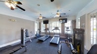 This fitness center offers a thoughtfully designed workout environment that balances functionality with comfort. The single-floor space is outfitted with a solid selection of cardio equipment, including multiple treadmills and an elliptical trainer, alongside strength and resistance machines to support a well-rounded fitness routine. The room is finished with crown molding and neutral-toned walls, lending a polished and welcoming aesthetic that sets it apart from a typical gym setting. Underfoot, dark carpeting covers the floor throughout the space, providing a cushioned surface suited for a variety of exercises. Multiple ceiling fans are evenly distributed across the ceiling, ensuring consistent air circulation during workouts. Two wall-mounted flat-screen televisions are positioned at opposite ends of the room, allowing exercisers to stay entertained from any station. Large windows along the exterior wall flood the space with natural light and frame views of the surrounding greenery, creating an open and airy atmosphere. Recessed lighting supplements the natural light, keeping the space bright and well-lit at all times. Overall, the fitness center presents a clean, comfortable, and fully equipped environment designed to meet a broad range of workout needs.