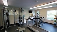 This fitness room is a well-equipped, single-floor exercise space designed to support a range of workout routines. The room features a dark, rubberized flooring that provides durability and comfort underfoot, while neutral-toned walls and fluorescent overhead lighting create a clean, functional environment. Natural light filters in through windows along one wall, adding brightness to the space.

Cardio equipment is a focal point of the room, including a treadmill and an elliptical machine positioned toward the center and rear of the space. A wall-mounted flat-screen television is positioned above the cardio area, allowing users to stay entertained during workouts. A dumbbell rack stocked with a range of free weights lines one side of the room, accompanied by a flat workout bench, making the space suitable for strength training. A cable machine provides additional resistance training options.

A standing fan offers supplemental air circulation, and a water cooler ensures hydration is conveniently accessible. A small waste bin is also present. The overall layout is efficiently organized to maximize functionality within a modest footprint, making it a practical amenity for everyday fitness use.