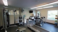 This fitness room is a well-equipped, single-floor exercise space designed to support a range of workout routines. The room features a dark, rubberized flooring that provides durability and comfort underfoot, while neutral-toned walls and fluorescent overhead lighting create a clean, functional environment. Natural light filters in through windows along one wall, adding brightness to the space.

Cardio equipment is a focal point of the room, including a treadmill and an elliptical machine positioned toward the center and rear of the space. A wall-mounted flat-screen television is positioned above the cardio area, allowing users to stay entertained during workouts. A dumbbell rack stocked with a range of free weights lines one side of the room, accompanied by a flat workout bench, making the space suitable for strength training. A cable machine provides additional resistance training options.

A standing fan offers supplemental air circulation, and a water cooler ensures hydration is conveniently accessible. A small waste bin is also present. The overall layout is efficiently organized to maximize functionality within a modest footprint, making it a practical amenity for everyday fitness use.