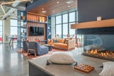 Located in Madison, WI, this contemporary amenity space occupies an upper floor and an expansive rooftop terrace, offering a well-appointed environment for gathering, working, and relaxing. The interior is defined by dramatic floor-to-ceiling windows that flood the space with natural light and frame sweeping views of the Madison skyline and the shimmering lake beyond. Polished concrete floors, exposed ductwork, and pendant lighting lend an industrial-modern aesthetic throughout.

The main interior area features a comfortable lounge with a built-in fireplace, wood-paneled shelving, and a large leather sofa — creating a warm, inviting retreat. Adjacent to the lounge, a full kitchenette is equipped with white cabinetry, warm wood open shelving, stainless steel appliances, and a retro red refrigerator that adds a playful accent. Multiple dining and café-style seating areas line the window wall, offering intimate spots to take in the panoramic views.

The rooftop terrace extends the experience outdoors, featuring bold red Adirondack chairs, a central fire pit, dual stainless steel grills, and ample dining seating — all set against unobstructed views of Lake Monona and the surrounding cityscape. Together, the interior and exterior areas create a dynamic, well-equipped space ideal for community and entertainment.