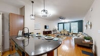 This well-appointed single-floor residence showcases a thoughtfully designed open-concept layout with warm hardwood flooring that flows seamlessly throughout the main living areas. At the heart of the home is a chef's kitchen featuring rich wood cabinetry, dark granite countertops, stainless steel appliances, and a large curved island with bar seating and built-in wine storage — all illuminated by stylish pendant lighting. The kitchen opens directly into a comfortable living area highlighted by an arched display niche, a wall-mounted TV, and a modern sputnik chandelier, creating an inviting atmosphere for entertaining. The formal dining room stands out with a bold teal accent wall, large windows that invite natural light, and contemporary ceiling lighting. The bedrooms offer plush carpeting, ample natural light through window treatments, and ceiling fans for year-round comfort. The primary bathroom is a spa-like retreat, complete with tile flooring, a soaking tub, a glass-enclosed walk-in shower, a generous double-sink vanity with dark granite counters, and warm wood cabinetry. A dedicated laundry room with a stacked washer and dryer and built-in shelving adds practical convenience to this polished and cohesive living space.