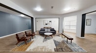 This single-floor communal space offers a well-designed, open-concept layout that blends modern aesthetics with functional comfort. The main lounge area is anchored by a charcoal gray sofa paired with mid-century modern wooden armchairs upholstered in rich brown leather, all arranged around a round white coffee table and a low wooden bench. A geometric patterned area rug in neutral earth tones grounds the seating arrangement, adding texture and visual warmth to the space. Abstract artwork adorns the white accent wall behind the sofa, complementing the clean, contemporary design sensibility. Warm wood-look luxury vinyl flooring extends throughout the entire space, providing a cohesive and durable foundation. The layout opens through wide, unobstructed doorways into adjacent corridor areas, which feature bold slate-blue accent walls that contrast with the crisp white trim and ceilings. These connecting hallways are spacious and well-lit, with flush-mount ceiling lights providing consistent, even illumination across all areas. A window with horizontal blinds allows natural light to filter into the lounge. The overall ambiance is polished and inviting, reflecting a thoughtfully curated design that balances modern style with practical, communal living needs.