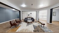 This single-floor communal space offers a well-designed, open-concept layout that blends modern aesthetics with functional comfort. The main lounge area is anchored by a charcoal gray sofa paired with mid-century modern wooden armchairs upholstered in rich brown leather, all arranged around a round white coffee table and a low wooden bench. A geometric patterned area rug in neutral earth tones grounds the seating arrangement, adding texture and visual warmth to the space. Abstract artwork adorns the white accent wall behind the sofa, complementing the clean, contemporary design sensibility. Warm wood-look luxury vinyl flooring extends throughout the entire space, providing a cohesive and durable foundation. The layout opens through wide, unobstructed doorways into adjacent corridor areas, which feature bold slate-blue accent walls that contrast with the crisp white trim and ceilings. These connecting hallways are spacious and well-lit, with flush-mount ceiling lights providing consistent, even illumination across all areas. A window with horizontal blinds allows natural light to filter into the lounge. The overall ambiance is polished and inviting, reflecting a thoughtfully curated design that balances modern style with practical, communal living needs.