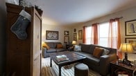 This single-floor residence offers a well-organized and functional layout designed for comfortable everyday living. The galley-style kitchen is efficiently arranged with warm honey-toned oak cabinetry lining both sides, providing generous storage space including upper and lower cabinets as well as above-cabinet shelving. The kitchen is equipped with a gas range, a full-size refrigerator, and a sink with a dish drying area. Stone-look tile flooring and overhead fluorescent lighting give the kitchen a practical, well-lit character. The kitchen flows naturally into a cozy dining area, where a ceiling fan light fixture casts a warm glow over a dark wood dining table and chairs. Decorative wall accents and warm-toned lighting add an intimate ambiance to the dining space. The living room is inviting and thoughtfully arranged, featuring a large sectional sofa and a loveseat in deep gray, accented with mustard and amber throw pillows that complement the warm color palette throughout. A rustic dark wood coffee table serves as a centerpiece, while framed artwork and maps adorn the walls, adding character and personality. Windows dressed with rich orange curtains allow natural light to filter in, completing the warm, lived-in feel of this comfortable residential space.