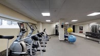 The fitness center is a single-floor space designed to support a well-rounded workout routine. The room features a row of cardio machines, including ellipticals and stationary bikes, arranged along one side to maximize floor space and ease of movement between equipment. A dedicated strength-training area is equipped with a multi-use cable machine, providing options for a variety of resistance exercises. For functional and flexibility training, the space includes a selection of stability balls in multiple sizes, stored neatly near a mirrored wall that enhances the sense of depth and allows users to monitor their form during workouts. The mirrored wall is a notable feature, reflecting both the equipment and the overhead lighting to brighten the room. The flooring consists of neutral, low-pile carpet suited for the space, while the drop-panel ceiling houses recessed flat lighting panels that provide consistent, even illumination throughout. The walls are finished in a neutral tone, contributing to a clean and focused atmosphere. Overall, the fitness center is practical and organized, offering residents or guests a comfortable and functional environment for their fitness needs.