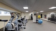 The fitness center is a single-floor space designed to support a well-rounded workout routine. The room features a row of cardio machines, including ellipticals and stationary bikes, arranged along one side to maximize floor space and ease of movement between equipment. A dedicated strength-training area is equipped with a multi-use cable machine, providing options for a variety of resistance exercises. For functional and flexibility training, the space includes a selection of stability balls in multiple sizes, stored neatly near a mirrored wall that enhances the sense of depth and allows users to monitor their form during workouts. The mirrored wall is a notable feature, reflecting both the equipment and the overhead lighting to brighten the room. The flooring consists of neutral, low-pile carpet suited for the space, while the drop-panel ceiling houses recessed flat lighting panels that provide consistent, even illumination throughout. The walls are finished in a neutral tone, contributing to a clean and focused atmosphere. Overall, the fitness center is practical and organized, offering residents or guests a comfortable and functional environment for their fitness needs.