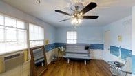 This single-room tanning space is designed with a distinct coastal ambiance, making it a unique and functional retreat. The room features warm hardwood floors that add a natural, inviting feel underfoot. A decorative ceiling fan with a light fixture is centrally mounted, providing both air circulation and ambient lighting to complement the space.

The most striking design element is the hand-painted ocean mural that wraps around the lower half of the walls, depicting a serene seascape complete with waves and sky. The upper walls are painted in a soft, pale blue that seamlessly complements the mural, reinforcing the beach-inspired theme throughout.

A horizontal tanning bed is positioned as the room's centerpiece, ready for use. A large, ornately framed floor mirror leans against one wall, adding a touch of elegance and practicality. A small side table and a portable fan are also present, offering convenience during sessions.

Two windows fitted with horizontal blinds allow for natural light control while maintaining privacy. Overall, the room delivers a purposeful, themed tanning environment with thoughtful decorative touches.