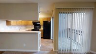 This single-floor space offers a practical and well-organized layout centered around an open kitchen and adjoining living area. The kitchen is equipped with warm-toned wood cabinetry providing ample storage, black appliances including a range with overhead microwave and a full-size refrigerator, and a built-in peninsula that serves as a natural divider between the kitchen and the living space. The peninsula offers additional counter space and provides a convenient area for casual dining or meal preparation. The kitchen features hard surface flooring, while the adjacent living area transitions to carpeting, subtly defining each zone. A sliding glass door fitted with vertical blinds allows natural light to filter into the space and opens to an outdoor balcony, offering views of the surrounding exterior. The neutral wall tones and standard ceiling height throughout contribute to a clean, functional atmosphere. The overall design prioritizes practicality, with a straightforward floor plan that connects the kitchen and living areas in a cohesive, easy-to-navigate arrangement.